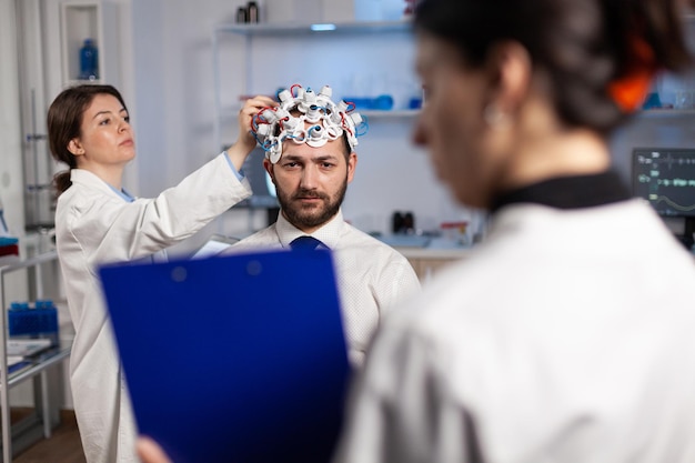 Foto facharzt für neurologe, der eine zwischenablage hält und dem patienten die neurologische behandlung erklärt, während die forscherin das eeg-headset anpasst. arzt überwacht die entwicklung der gehirnaktivität