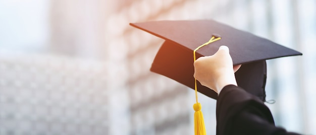 fechar mão de pós-graduação segurar mostrar chapéu no prédio da escola de fundo. Tiro do chapéu de formatura durante o curso universitário de início, conceito de aprendizagem de sucesso de estudante de educação.