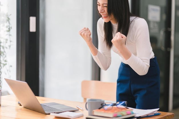 Foto feliz e entusiasmada, uma jovem asiática, estudante ou funcionária, celebra a realização de seu objetivo de ganhar uma competição on-line ou receber boas notícias por e-mail, levantando as mãos em euforia.