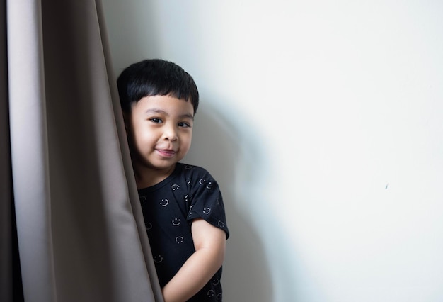 Feliz menino asiático de três anos posando sobre cortinas marrons e fundo de paredes brancas