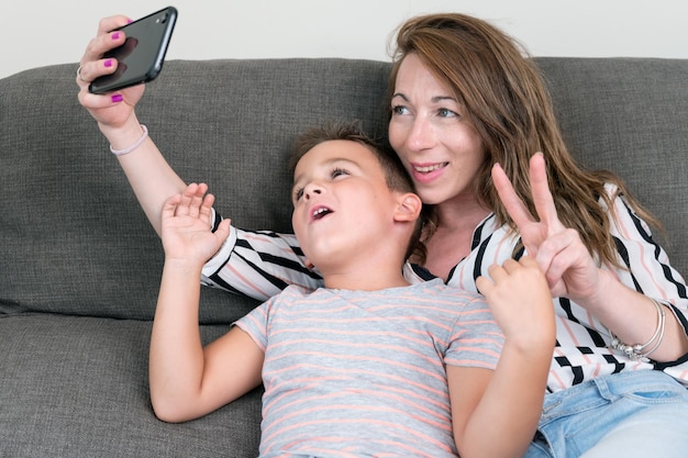 Fröhliche Mutter, die ein Smartphone hält, das Selfie mit ihrem kleinen Sohn macht, der Siegeszeichen zeigt