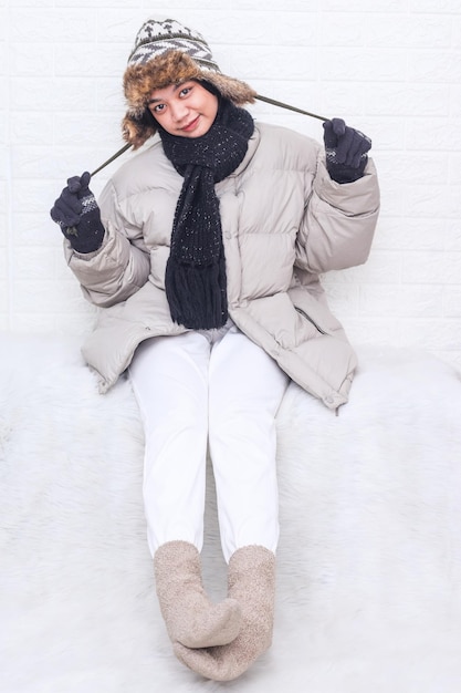 Fröhliches Mädchen mit Pufferjacke Pulloverbeanie Hutschnur sitzt im warmen Raum Wintermode