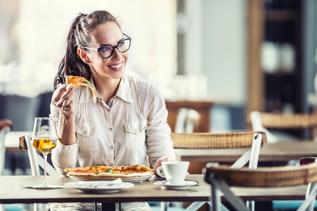 Garota bonita de óculos se senta em um restaurante segurando uma fatia de pizza na mão com um copo de vinho sorrindo