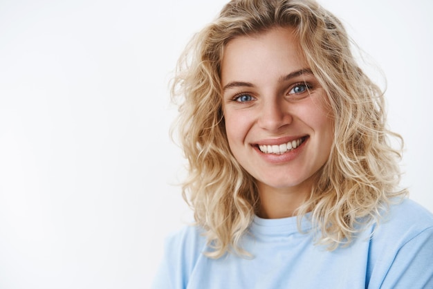 Garota com sorriso sincero fofo rindo e se divertindo olhando amigável e encantado com a câmera com profundos olhos azuis em pé alegremente sobre fundo branco em camiseta rindo paquerando