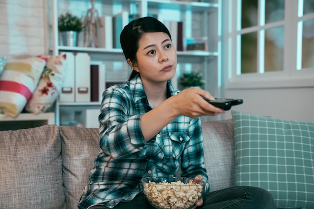 Foto garota entediada assistindo filme chato no sofá no quarto escuro. mulher asiática sentada no sofá comendo pipoca e zapping mudando de canal na tv pelo controlador. mulher fica acordada até tarde olhando programa de monitor à noite