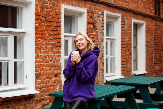 Glückliche blonde Frau im violetten Hoodie posiert im Fast-Food-Café im Freien und hält Kaffee zum Mitnehmen.