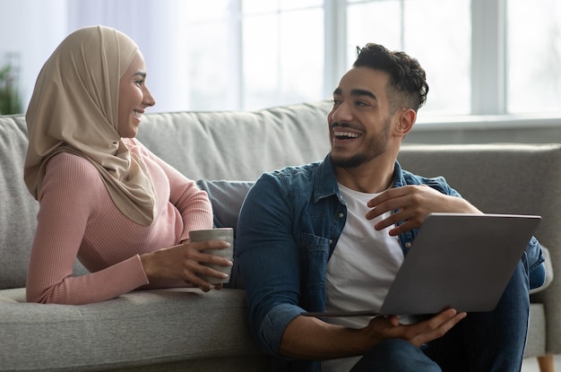 Glücklicher muslimischer bärtiger Mann und Frau im Hijab, die online Möbel auswählen, einen modernen Laptop benutzen, Tee trinken und sich unterhalten, ein gemeinsames Wochenende planen, Inneneinrichtung, Kopienraum