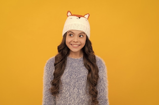 Glückliches Kind mit langen Haaren im Hut auf gelbem Hintergrund im Winter