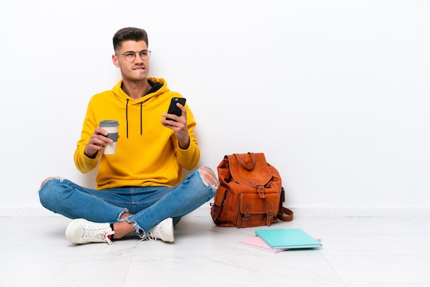 Homem caucasiano jovem estudante sentado no chão isolado no fundo branco segurando café para levar e um celular enquanto pensava em algo