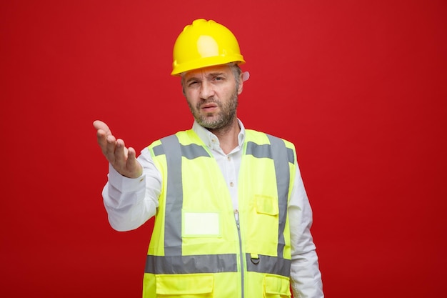 Homem construtor em uniforme de construção e capacete de segurança olhando para a câmera confuso e descontente, levantando o braço em desagrado em pé sobre fundo vermelho