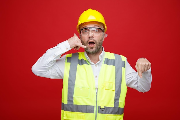 Homem construtor em uniforme de construção e capacete de segurança usando óculos de segurança olhando para a câmera espantado e surpreso fazendo me chamar de gesto apontando com o dedo indicador para baixo em pé sobre fundo vermelho