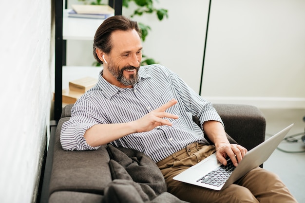 Homem de meia idade positivo e amigável sorrindo e acenando com a mão enquanto usa um laptop moderno e fones de ouvido sem fio durante a videochamada