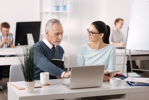 Homem encantador vestindo casaquinho azul usando laptop enquanto mostra novas mudanças no trabalho