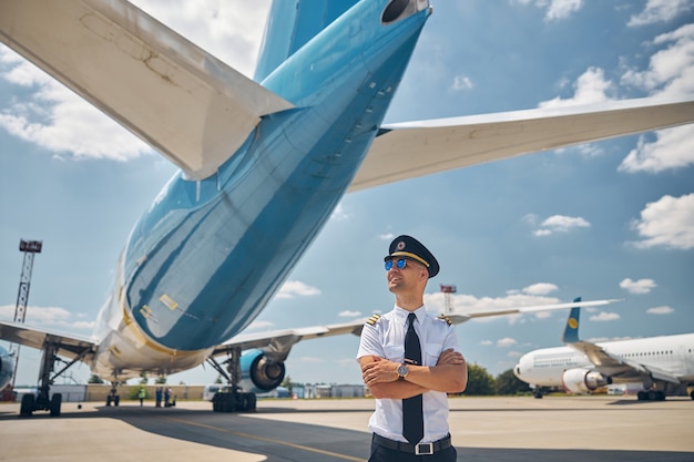 Foto hübscher junger mann, der die arme kreuzt und lächelt, während er im freien mit flugzeugen und himmel im hintergrund steht