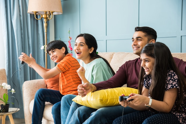 Indische junge vierköpfige Familie, die Videospiele mit Controller oder Joystick spielt, während sie auf dem Sofa sitzt sitting