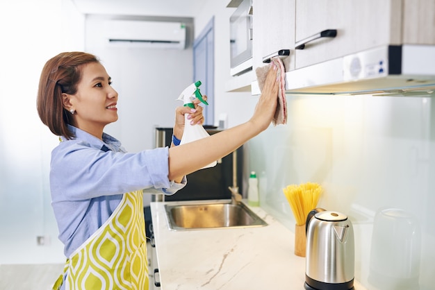 Foto jovem alegre dona de casa vietnamita limpando armários de cozinha com spray desinfetante