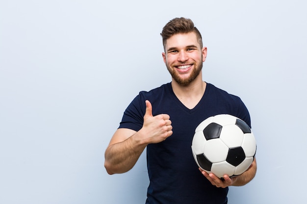 Jovem homem caucasiano segurando uma bola de futebol, sorrindo e levantando o polegar