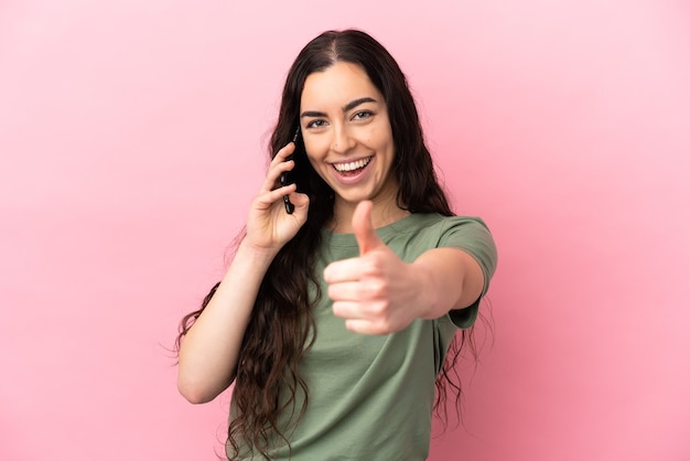 Jovem mulher caucasiana isolada em um fundo rosa, conversando com o celular enquanto faz o polegar para cima