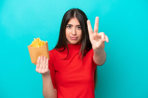 Jovem mulher caucasiana pegando batatas fritas isoladas em fundo azul sorrindo e mostrando sinal de vitória