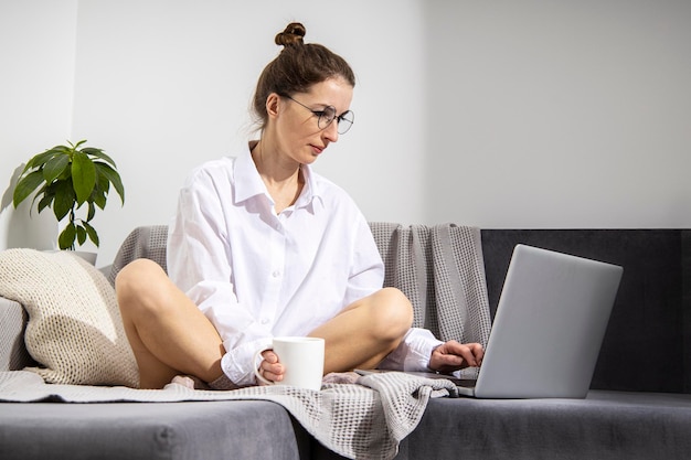 Jovem mulher trabalhando com um laptop com uma xícara de café sentada em um sofá