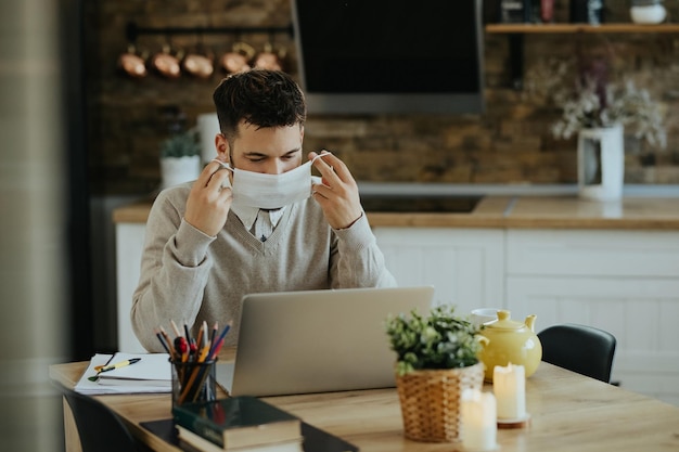 Jovem trabalhador autônomo usando máscara facial enquanto trabalhava no laptop em casa