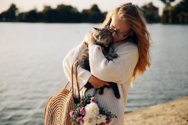 Junge blonde Frau mit Brille hält eine Katzenrasse Scottish Straight Walk mit Haustier in der Nähe des Sees bei Sonnenuntergang am Strand im Frühjahr