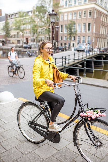 Junge Frau im gelben Regenmantel mit Tasche und Blumen mit dem Fahrrad in Amsterdam City