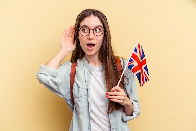 Junge kaukasische Frau, die Englisch isoliert auf gelbem Hintergrund lernt und versucht, einen Klatsch zu hören.