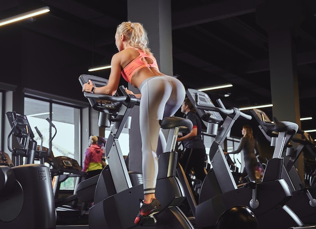 Junge schöne blonde Frau im Sportbekleidungstraining auf einem Heimtrainer in einem modernen Fitnesscenter.