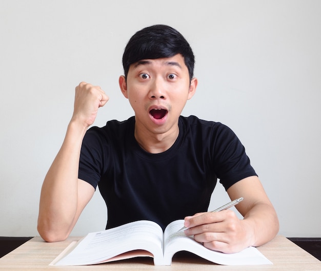 Junger asiatischer Mann fühlt sich schockiert aufgeregt Gesicht und Faust mit dem Buch auf dem Schreibtisch Blick in die Kamera