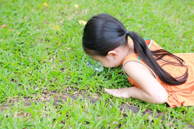 Kleines asiatisches Kindermädchen mit Lupe auf Garten des grünen Grases. Nahaufnahmenatur als Forscher.