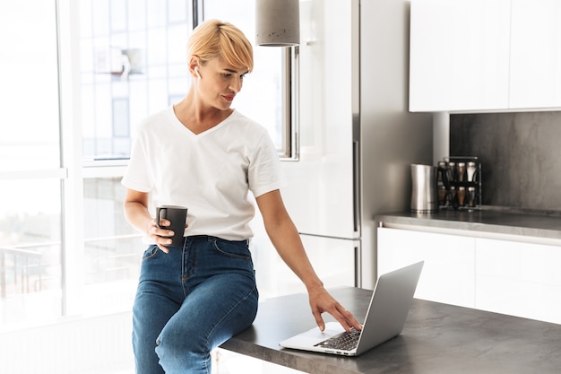 Lächelnde Frau lässig gekleidet mit Laptop-Computer beim Sitzen auf einer Küche, Kopfhörer tragend