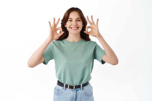 Lächelnde süße junge Frau im T-Shirt zeigt ihre Unterstützung, macht okayzeichen und nickt zustimmend, stimmt zu oder genehmigt, empfiehlt Produkt, steht gegen weiße Wand