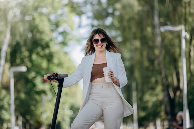 Foto linda jovem de terno branco e óculos de sol em pé com sua scooter elétrica no parque da cidade e bebendo café