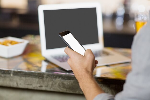 Mann, der Handy mit Laptop auf Tisch am Bartheke benutzt