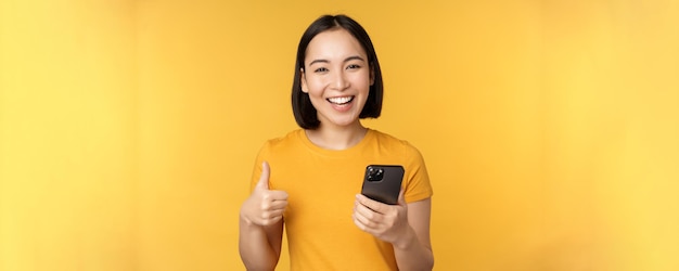 Menina asiática sorridente feliz segurando o celular e mostrando os polegares para cima recomendando o aplicativo no smartphone sobre fundo amarelo