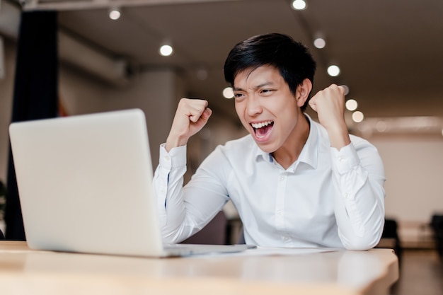 Foto millenial homem de negócios asiáticos no escritório com laptop feliz com sua vitória