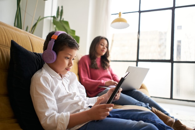 Millennial-Mutter, die mit ihrem kleinen Sohn von zu Hause aus arbeitet, das Kind spielt mit einem Tablet-Konzept der Familie ...