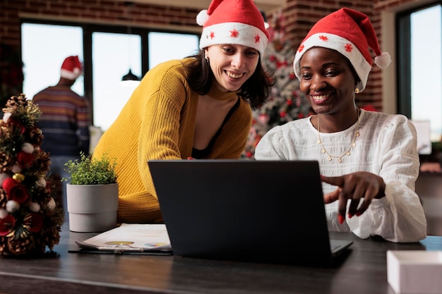 Mitarbeiter, die im festlichen Firmenbüro Teamarbeit leisten und am Laptop arbeiten, um ein Projekt in einem Raum zu planen, der mit Weihnachtsschmuck und Lichtern gefüllt ist. Menschen im Bürojob mit Weihnachtsbaum und Dekorationen.