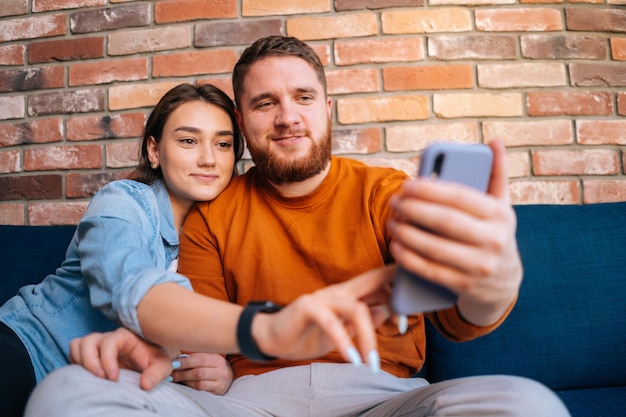 Mittelaufnahme Porträt eines glücklichen jungen Paares, das online per Webcam per Videoanruf auf dem Handy chattet und zusammen auf einem bequemen, weichen Sofa im gemütlichen Wohnzimmer sitzt. Konzept des Haushaltslebens.