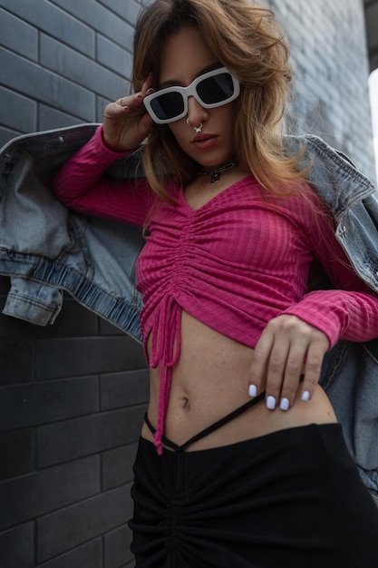 Moda sexy linda mulher ruiva hipster em streetwear elegante com jaqueta jeans rosa e saia preta fica e usa um óculos de sol legal branco perto de uma parede de tijolos pretos na cidade