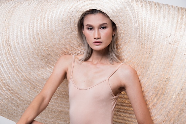 Foto mode junge dünne asiatische frau tan haut augen schön bilden mode rosa kleid posiert attraktiven glam-look sehr großen strohhut. studiobeleuchtung aus weißem hintergrund, sommerstrand heiße sonne