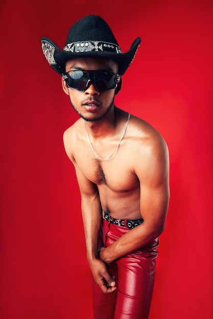 Modischer Mann und Cowboyhut, Sonnenbrille und hemdlose Kleidung und Gen Z isoliert auf rotem Hintergrund. Selbstbewusstes Model, cool und Lederhose mit Brillenaccessoire, westliche Ästhetik in einem Studio