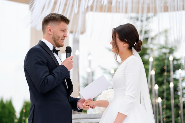 Momento de ternura de vista lateral no noivo da cerimônia de casamento fala um juramento de amor para a linda noiva Casal amoroso homem e mulher tendo cerimônia de casamento ao ar livre