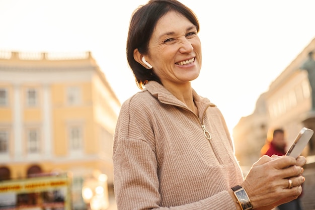 Foto mulher adulta caucasiana sorridente recebendo mensagem agradável de parentes no telefone enquanto está ao ar livre conceito de emoções positivas