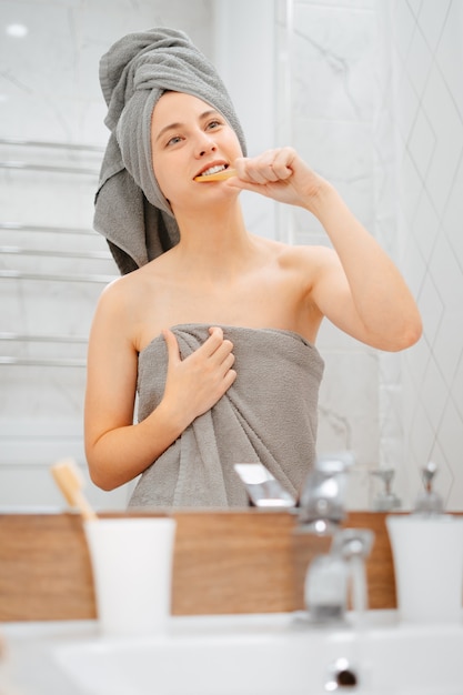 Mulher depois do banho com uma toalha cinza escova os dentes com uma escova de madeira com pasta clareadora