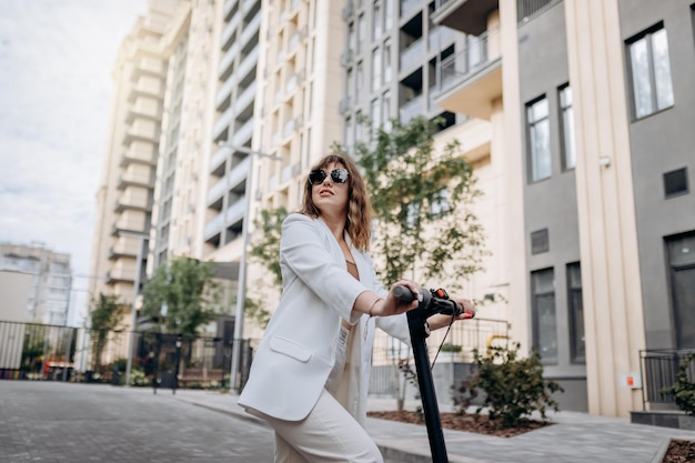 Foto mulher jovem e bonita em óculos de sol e terno branco em pé em sua scooter elétrica perto de um edifício moderno e desviar o olhar