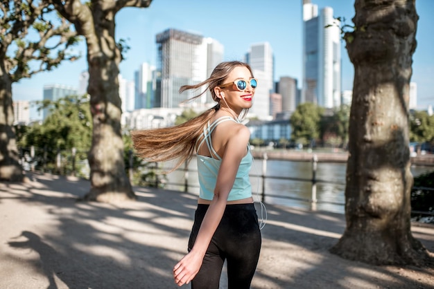 Foto mulher jovem em roupas esportivas, andando e olhando para o parque com arranha-céus ao fundo na cidade de frankfurt