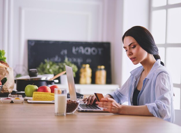 Mulher sorridente fazendo compras online usando computador e cartão de crédito na cozinha