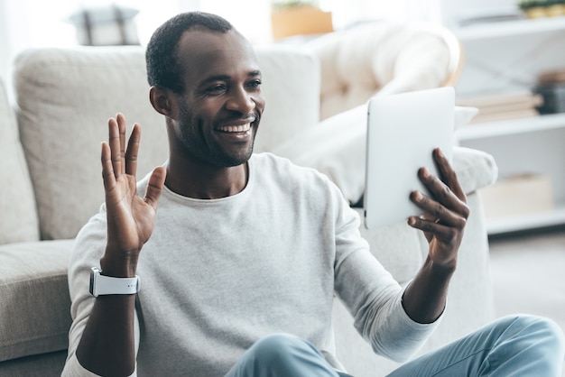 Olá! Africano jovem bonito olhando para seu tablet digital e sorrindo enquanto está sentado no tapete em casa